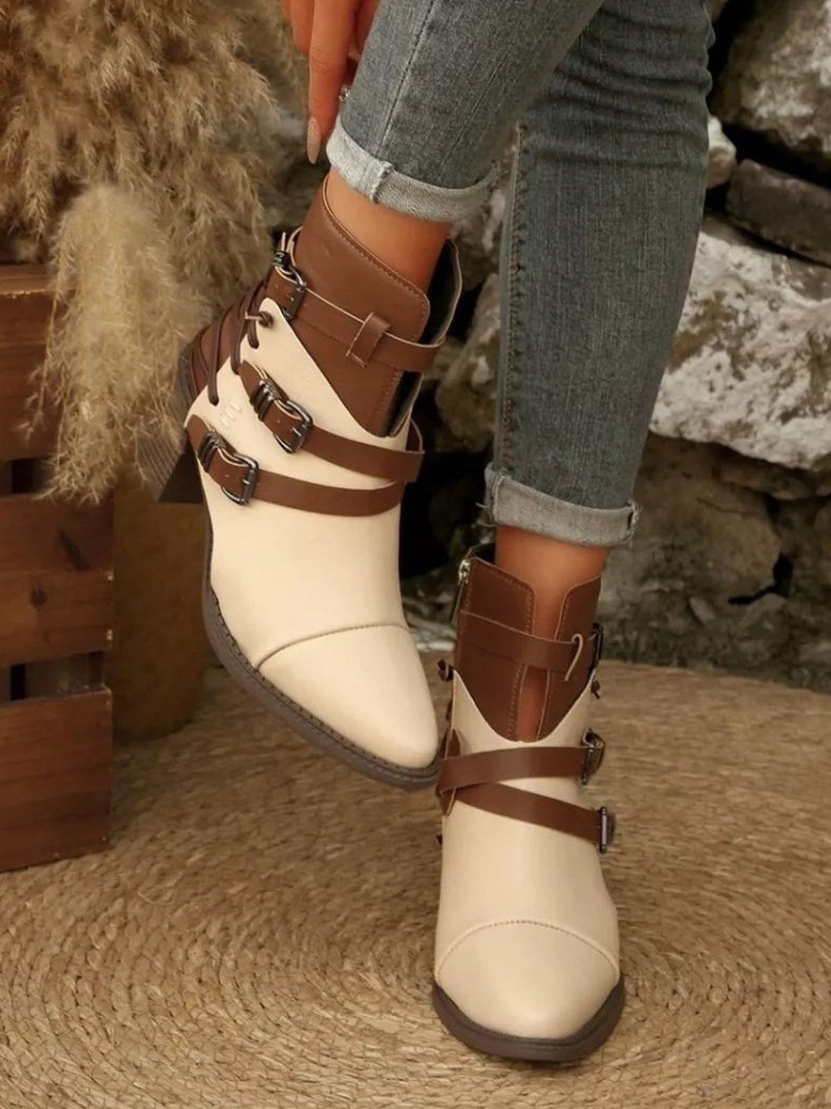 A model wears cream and brown point-toe ankle boots with multiple buckle straps, mid block heels, and contrasting panels, posed with one foot lifted.