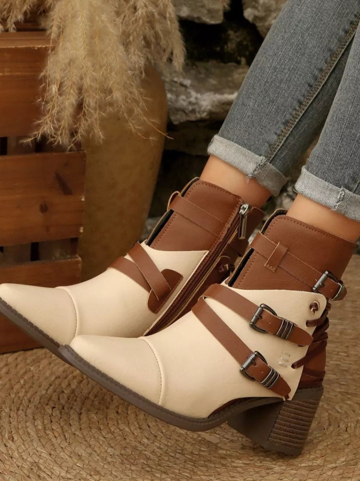 A model crosses her legs to showcase cream and brown point-toe ankle boots with multiple buckle straps, side zippers, and sturdy mid-height block heels.