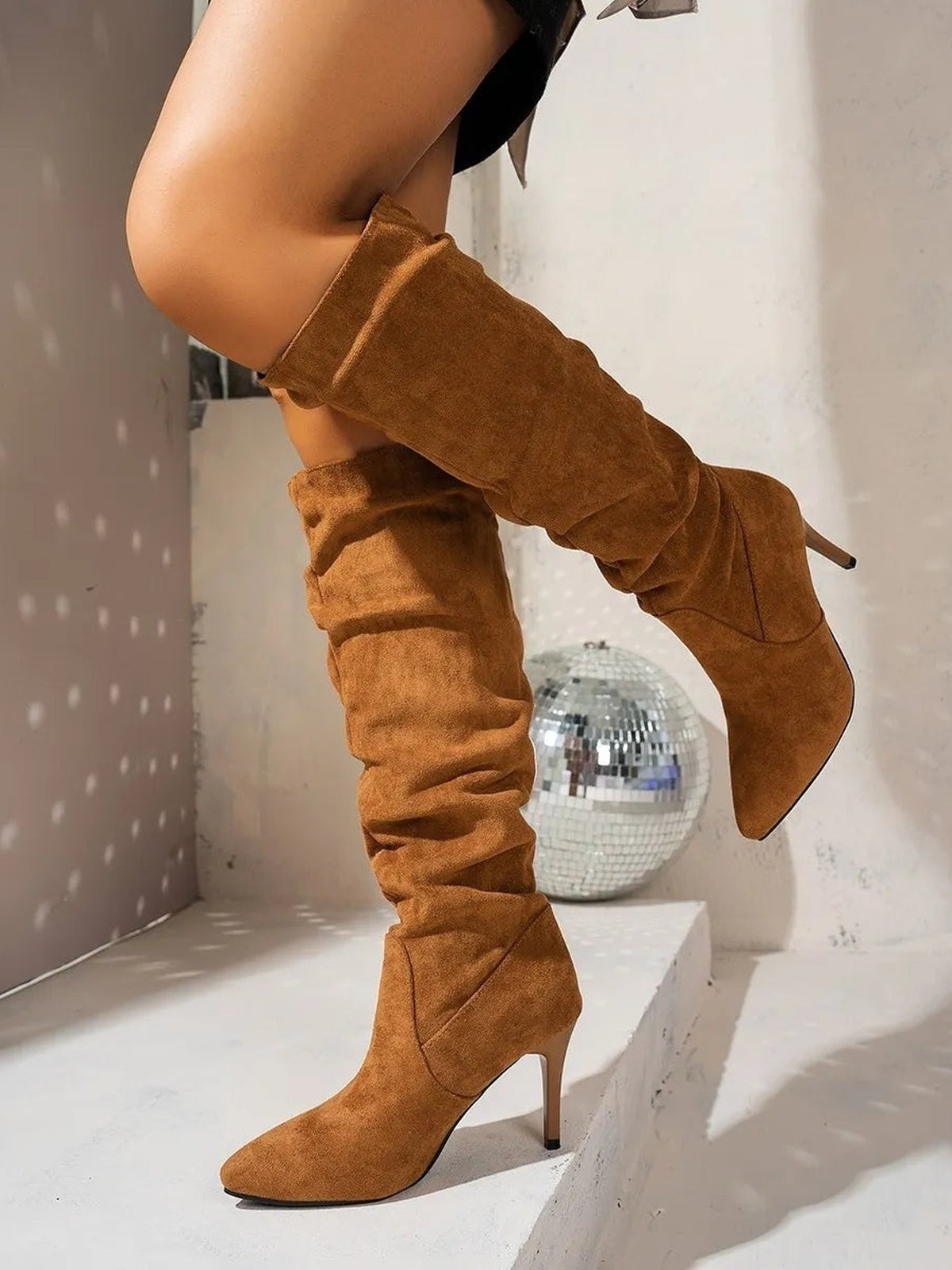 A model poses in camel suede point-toe stiletto boots with a slouchy over-the-knee silhouette, folded shaft, and slim high heels.