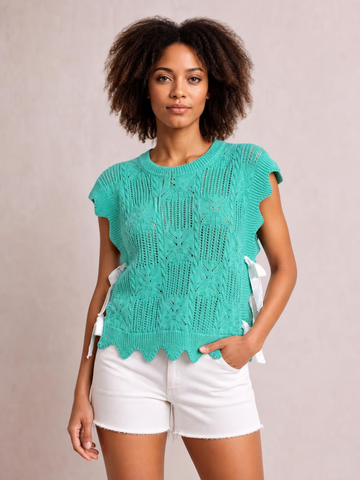 Model wearing a turquoise pointelle knit sweater with patterned openwork, short sleeves, scalloped trim, and a relaxed silhouette, one hand in her pocket.