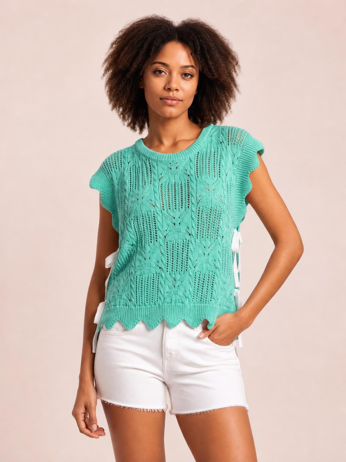 A model poses in a turquoise pointelle knit sweater with openwork patterning, short sleeves, side ties, relaxed silhouette, and scalloped hem trim.