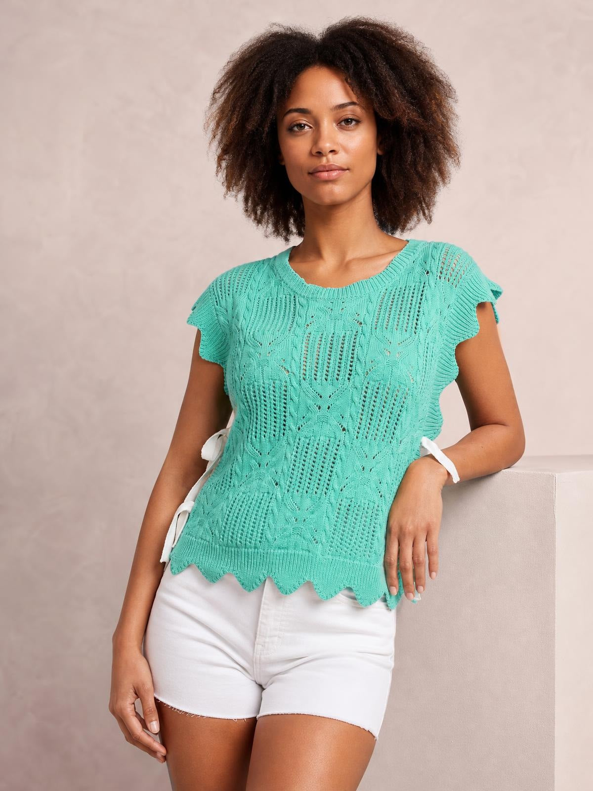 A model leans against a ledge wearing a turquoise pointelle knit patterned sweater with short sleeves and scalloped trim at the hem.