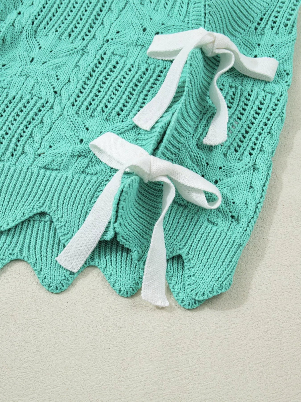 Close-up of a turquoise pointelle knit sweater with patterned openwork, ribbed sections, scalloped trim, and white tie details.