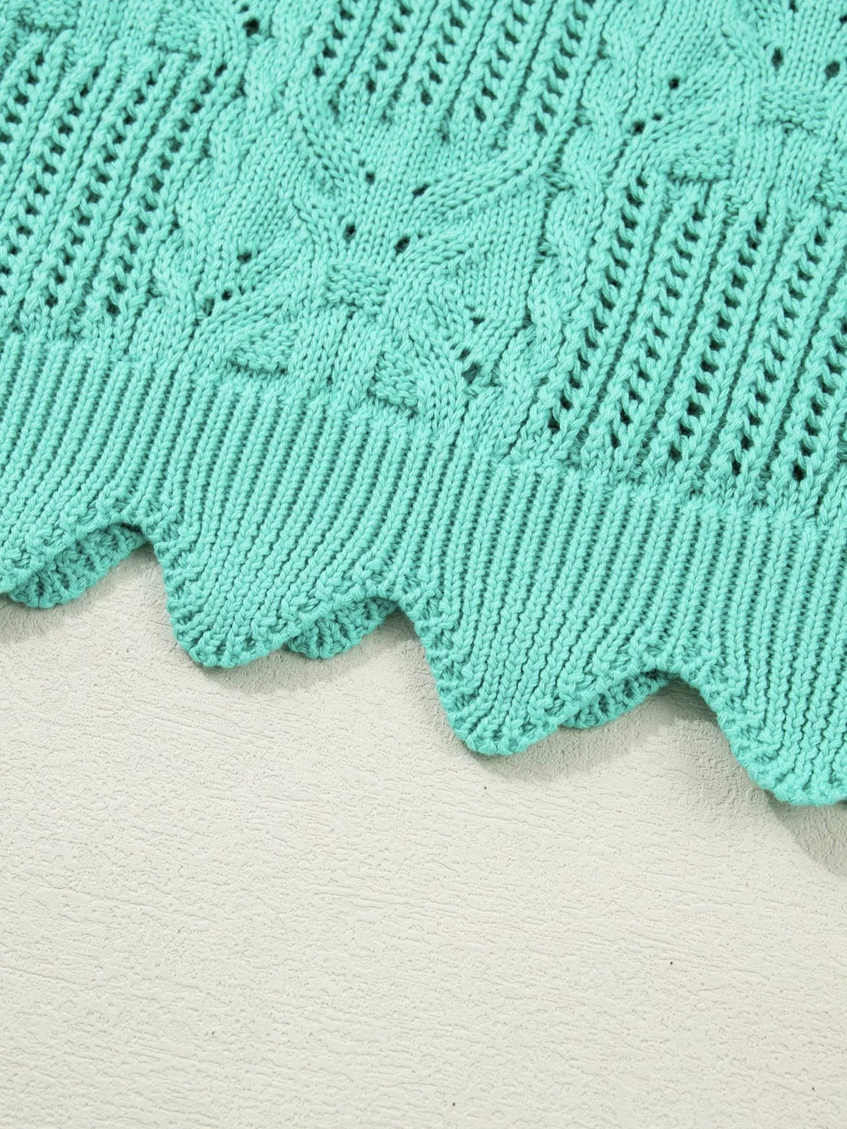 Close-up of a turquoise pointelle knit sweater showing patterned openwork texture, ribbed construction, and a scalloped trim hem.