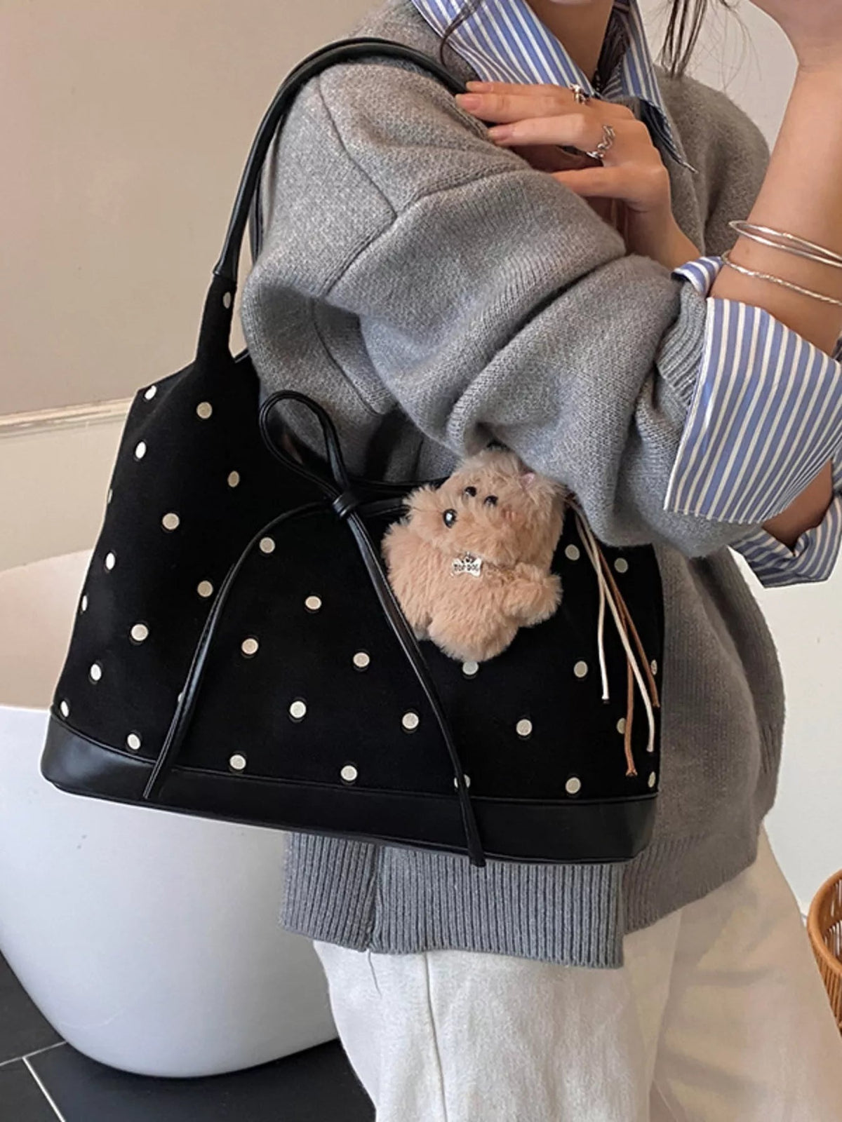 A model holds a medium black PU leather polka dot bow shoulder bag against her shoulder, showing its structured silhouette and long strap over a gray knit sweater.