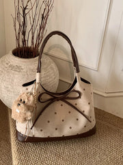 A medium cream PU leather shoulder bag with brown polka dots, contrast trim, rounded handles, a structured silhouette, and a large bow front detail.