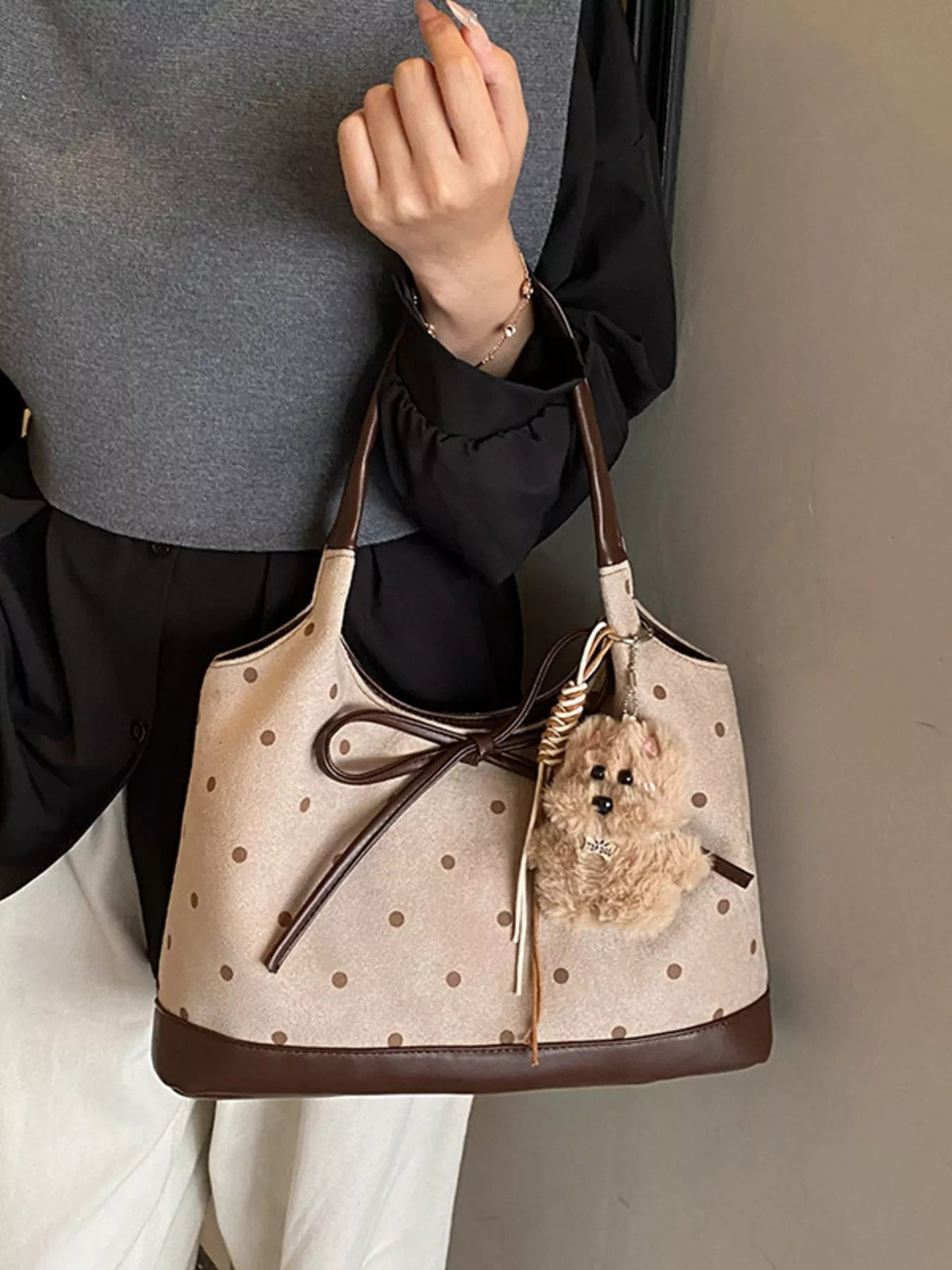 A model carries a medium beige PU leather shoulder bag with brown polka dots, a structured bucket silhouette, contrast trim, and a large bow detail.