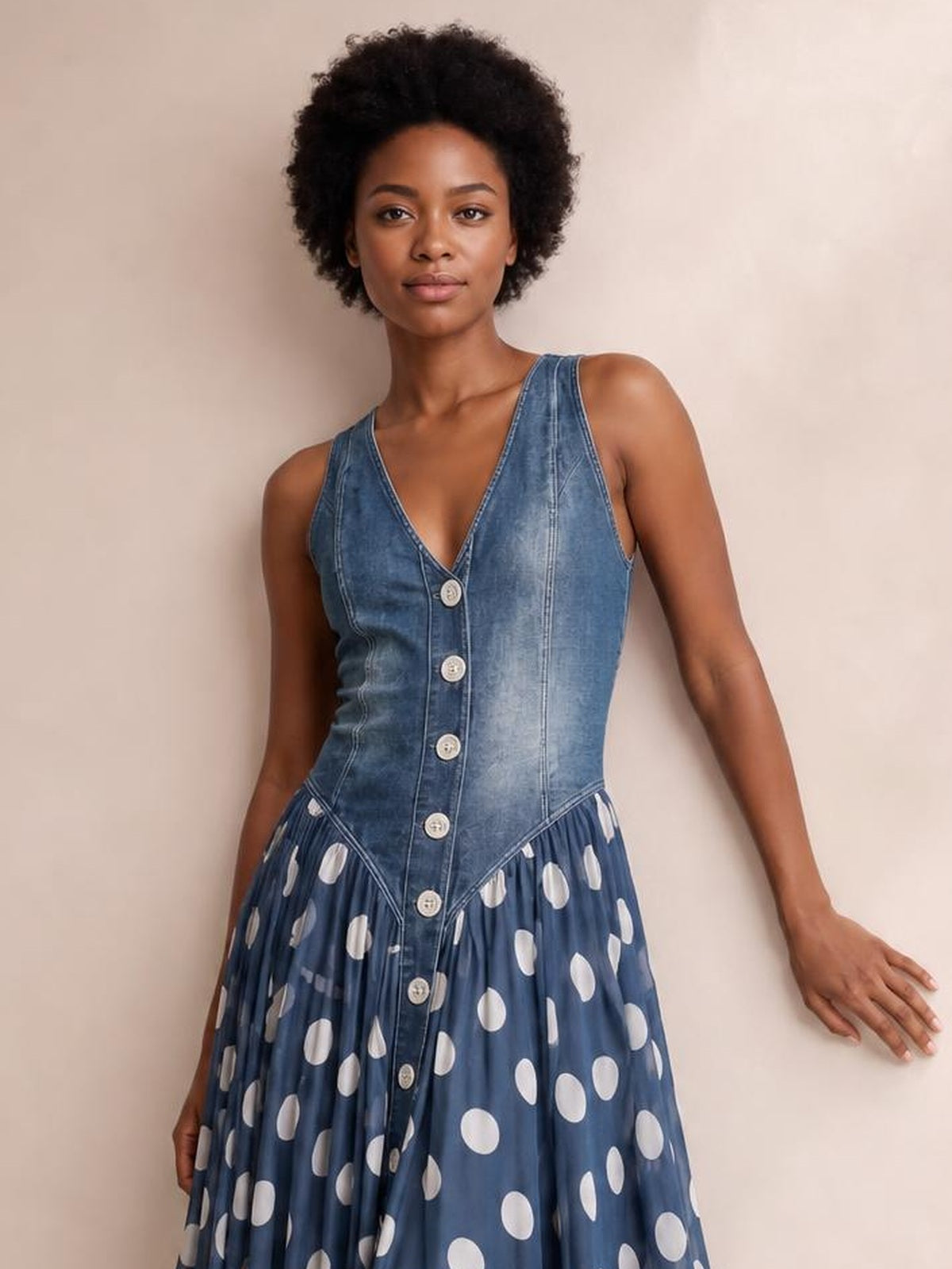 Model poses with one hand against the wall, wearing a blue denim sleeveless maxi dress with a V-neck bodice, button front, fitted seams, and gathered polka dot skirt.