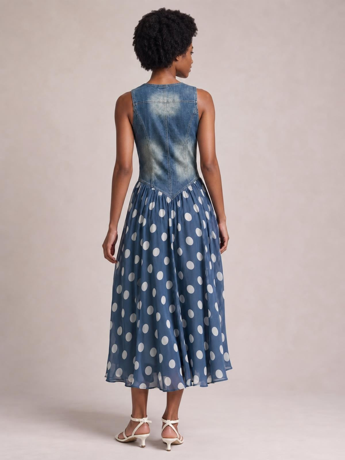 A model poses from the back in a sleeveless blue denim bodice dress with seamed construction, fitted waist, and a flowing polka dot maxi skirt.