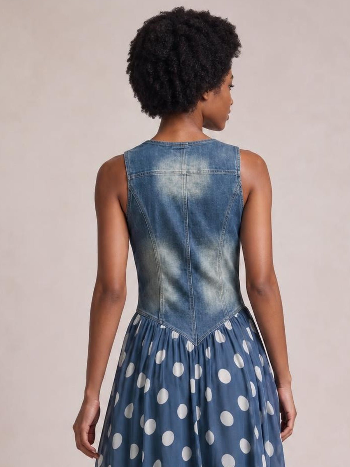 Model shown from the back in a sleeveless blue washed denim bodice dress with a fitted seamed construction and gathered polka dot maxi skirt.