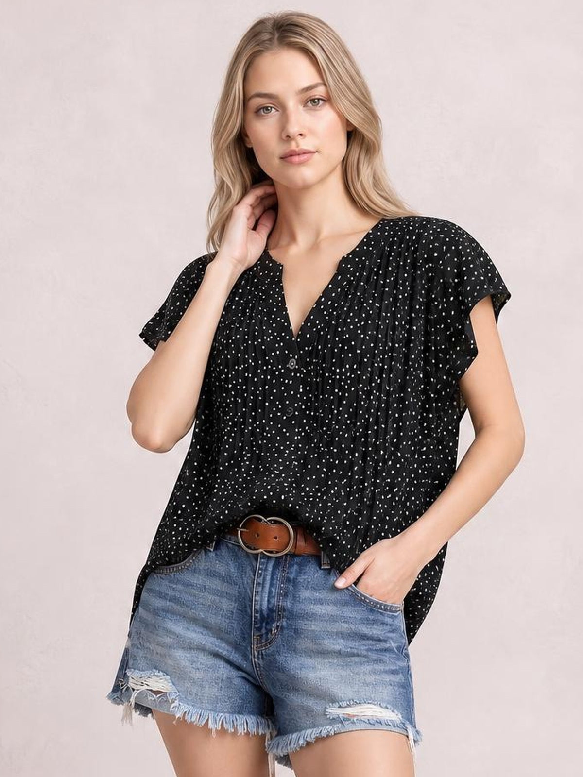 Model wears a black polka dot blouse with a notched neckline, ruffled cap sleeves, ruched gathers, and a relaxed, flowy silhouette.