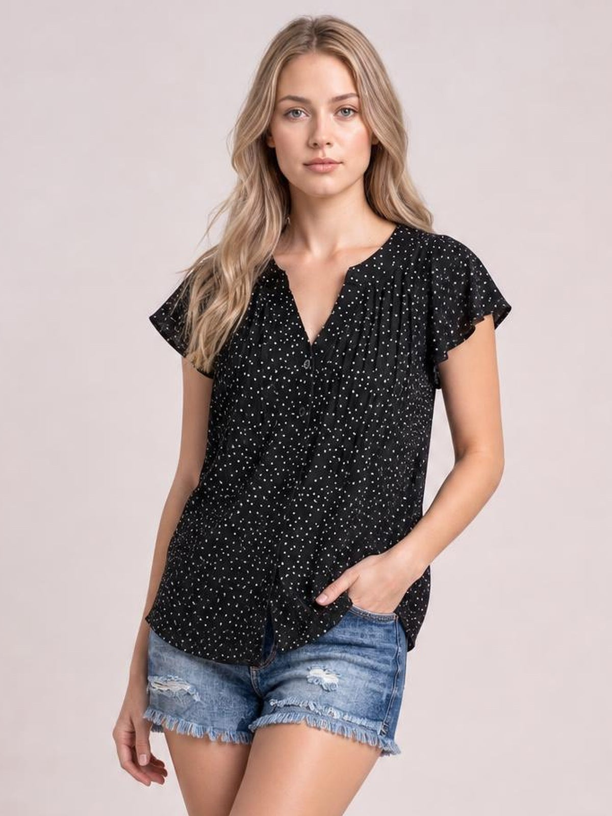 Model wears a black polka dot blouse with a notched neckline, ruffled cap sleeves, ruched front, button detail, and a relaxed silhouette.