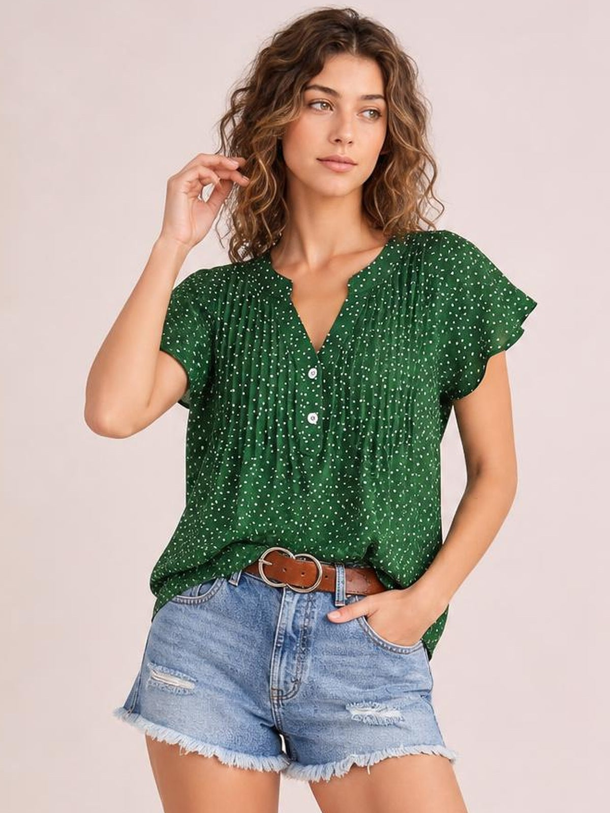 Model poses with one hand in her pocket wearing a green polka dot blouse with a notched neckline, ruched front, button placket, and ruffled cap sleeves.
