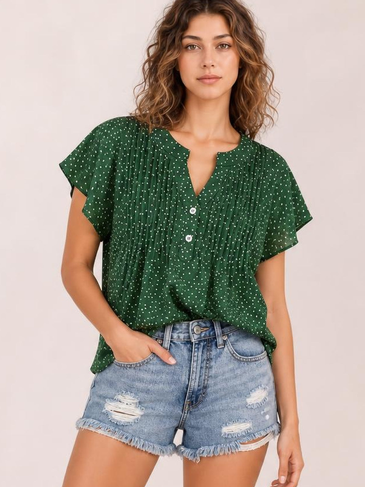 Model poses in a relaxed green polka dot blouse with a notched neckline, ruffled cap sleeves, ruched front, button closure, and loose silhouette.