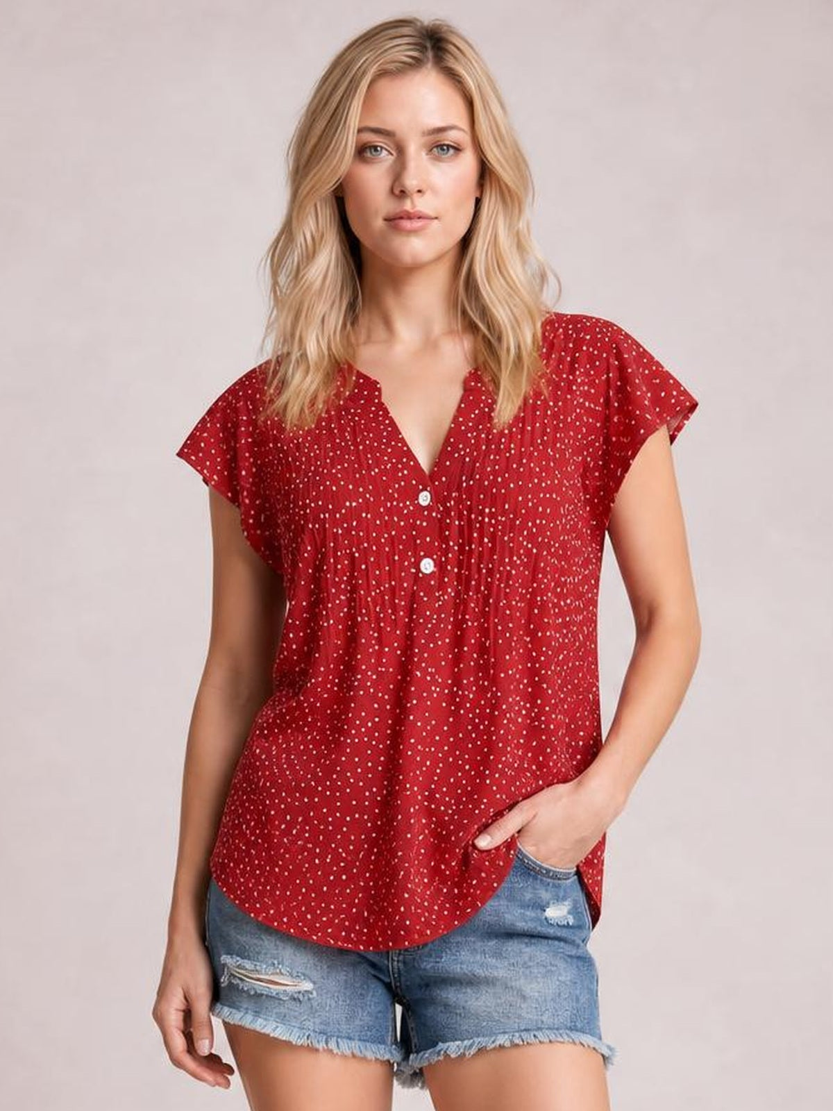 A model stands in a red polka dot blouse with a notched neckline, ruffled cap sleeves, button front, ruched detailing, and a relaxed curved hem.