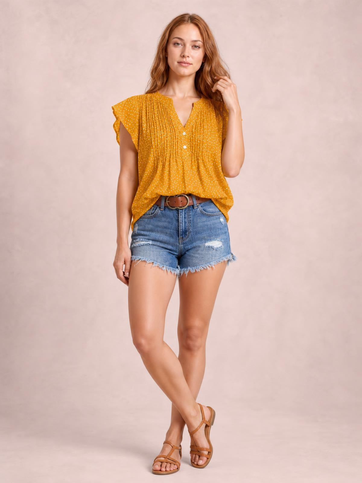 Model wears a mustard polka dot blouse with a notched V-neck, button front, ruched pleating, ruffled cap sleeves, and a relaxed flowing silhouette.