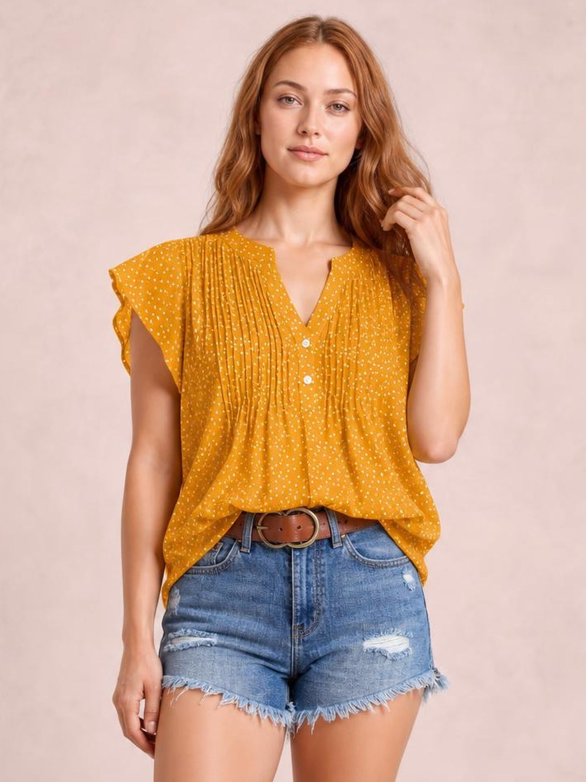 Model wearing a mustard polka dot blouse with a notched neckline, ruffled cap sleeves, ruched pleats, and a relaxed silhouette while touching her hair.
