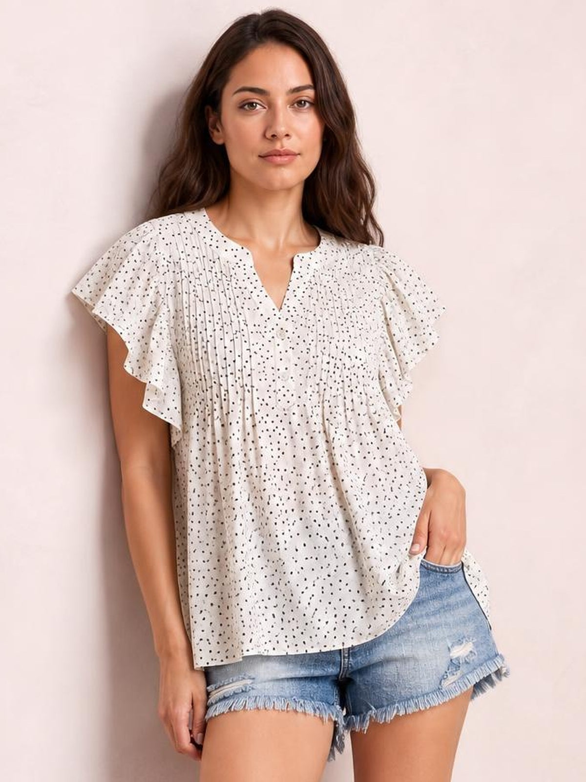 Model wears a white polka dot blouse with a notched neckline, ruched front, ruffled cap sleeves, and a relaxed silhouette, posing with one hand in her shorts pocket.