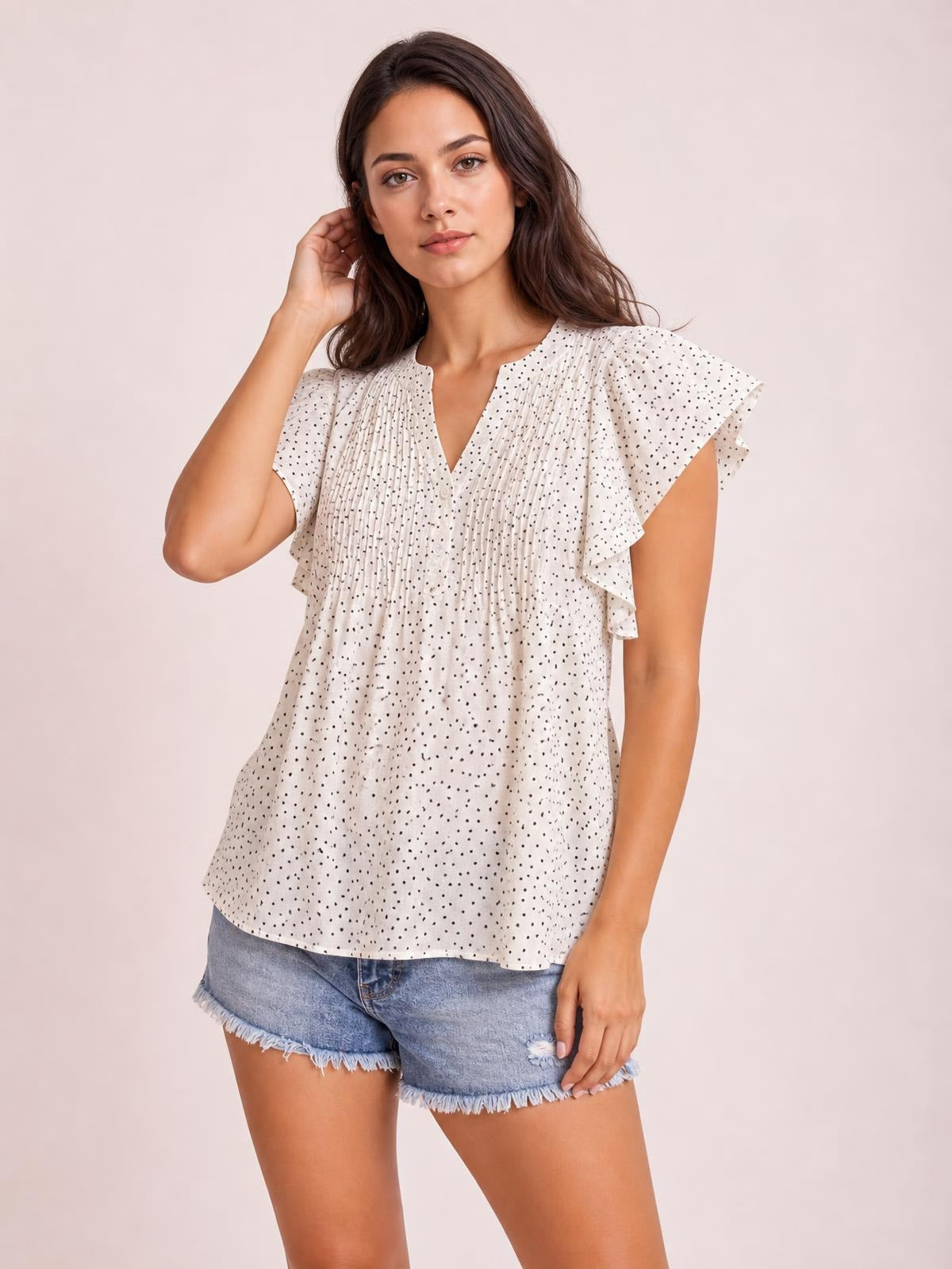 A model poses in a white polka dot blouse with a notched neckline, ruched front, ruffled cap sleeves, and a relaxed, flowing silhouette.