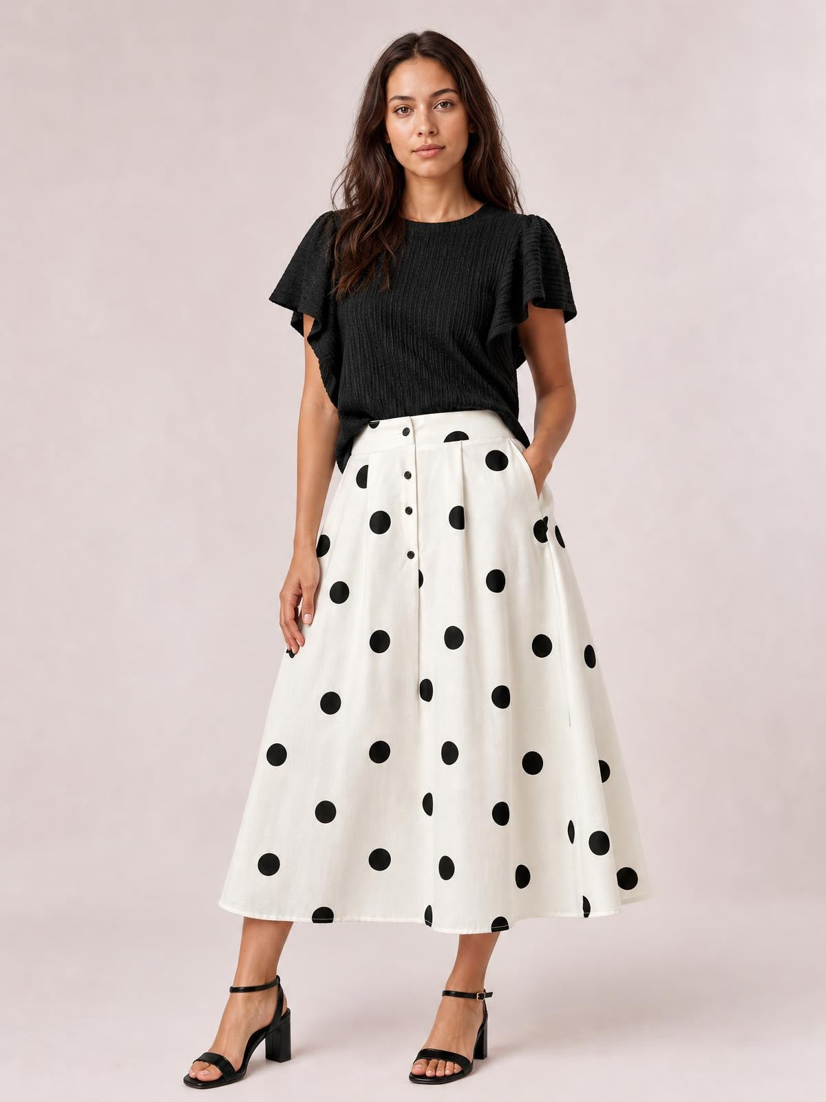 A model stands with one hand in a pocket, wearing a white Polka Dot Skirt with black spots, button-front detailing, and a flared midi silhouette.