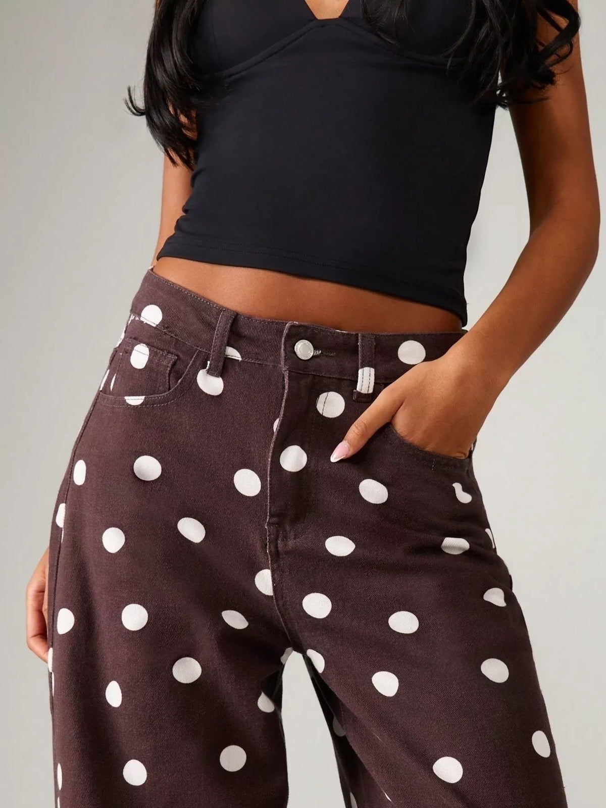 Model wears dark brown polka dot wide-leg jeans with a button fly, belt loops, front pockets, and a relaxed silhouette, posing with one hand in a pocket.