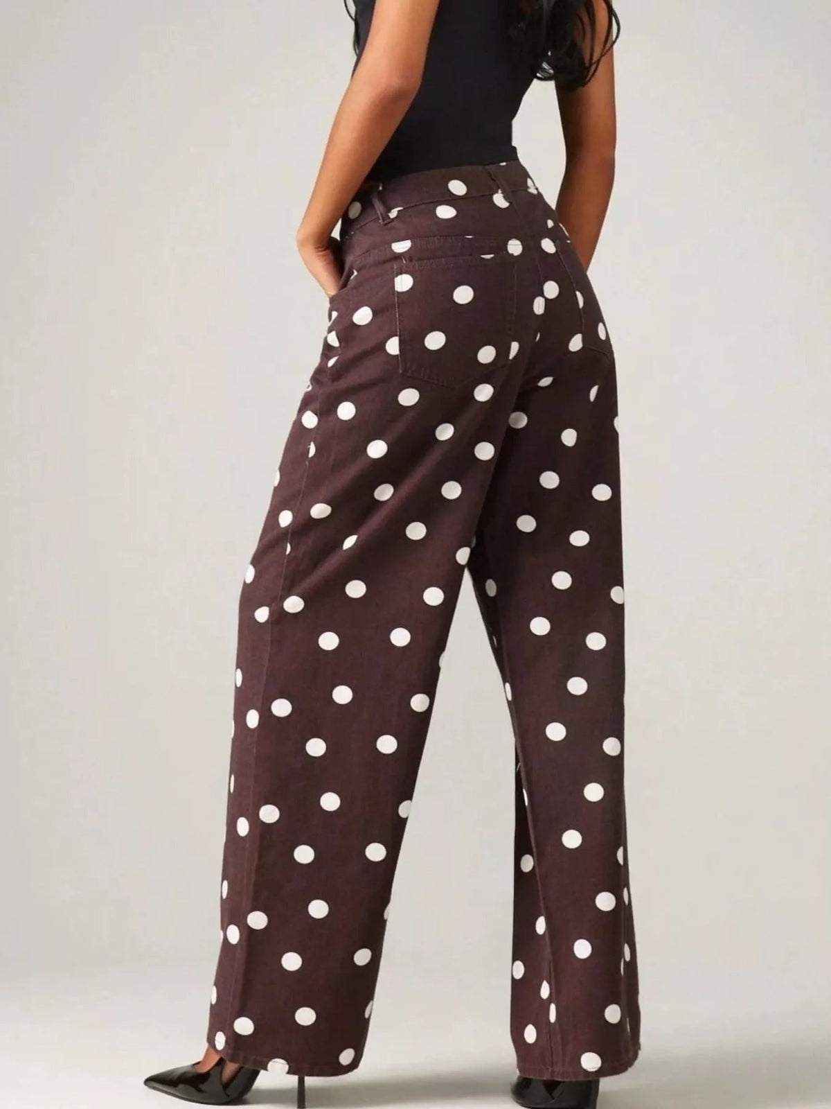 Model poses from the back with hands in the pockets of dark brown polka dot wide-leg jeans, showing a high waistband and relaxed full-length silhouette.