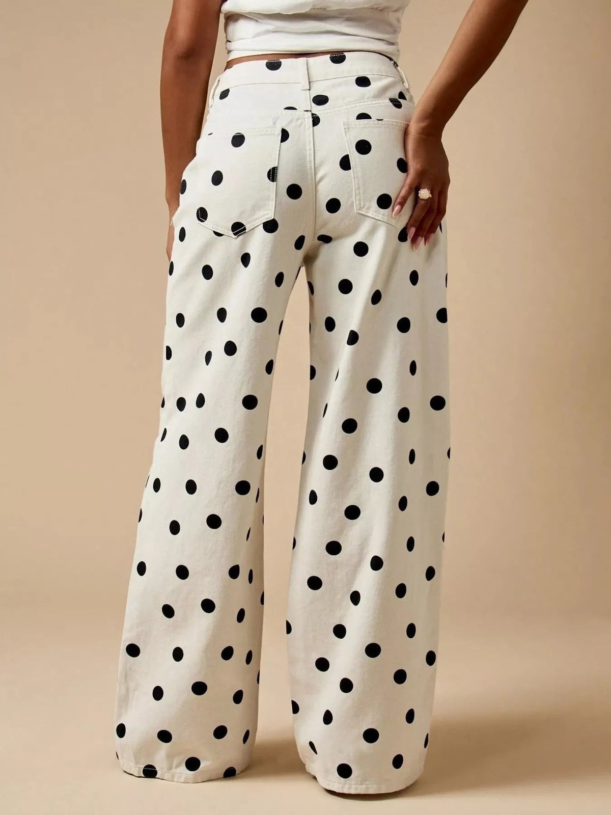 A model wears cream polka dot wide-leg jeans with black dots, a structured waistband, rear patch pockets, and a relaxed full-length silhouette.