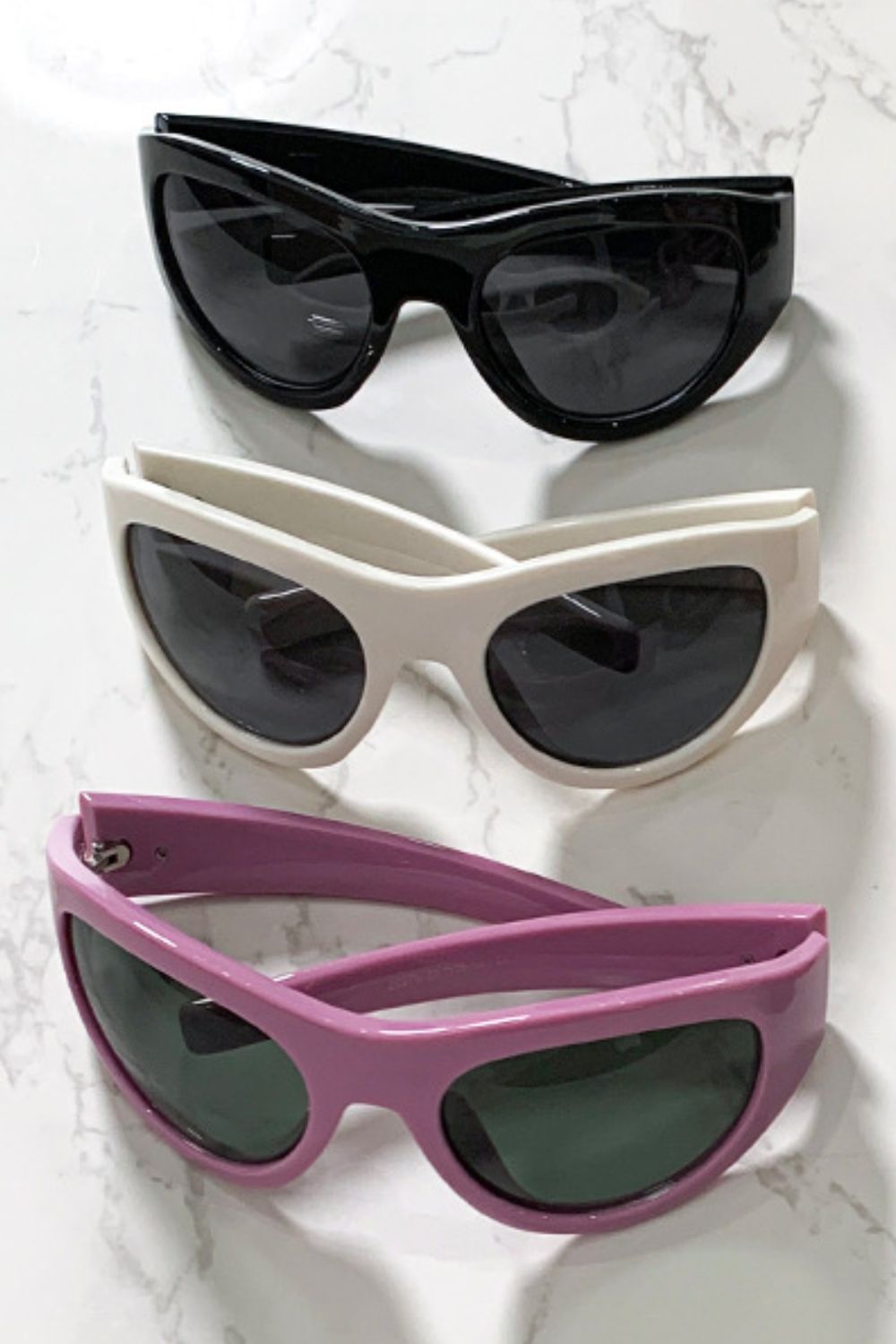 Three pairs of polycarbonate cat-eye sunglasses in black, ivory, and pink feature rounded dark lenses, softly upswept corners, and sculpted frames with tapered temples.