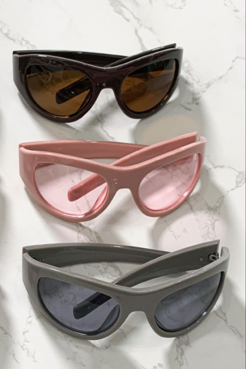 Three pairs of polycarbonate cat-eye sunglasses feature softly upswept frames, rounded lenses, tapered temples, and glossy black, pink, and gray colorways.