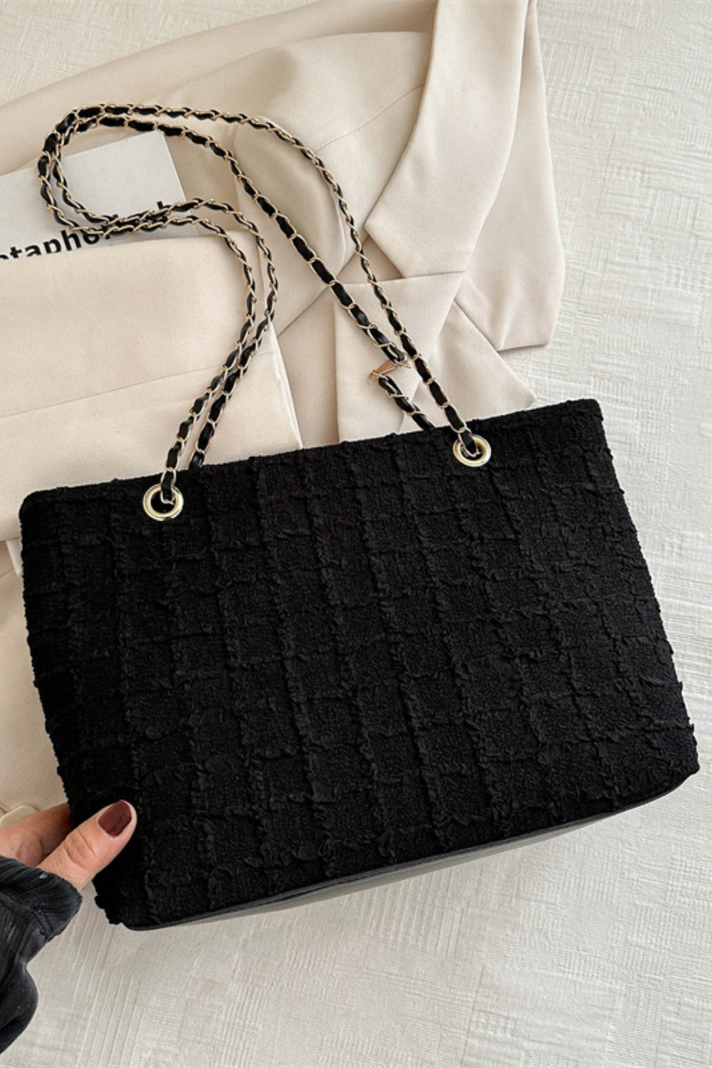 A hand displays a medium black polyester tote with a tactile textured surface, softly structured silhouette, and linked chain handles attached with gold-tone rings.