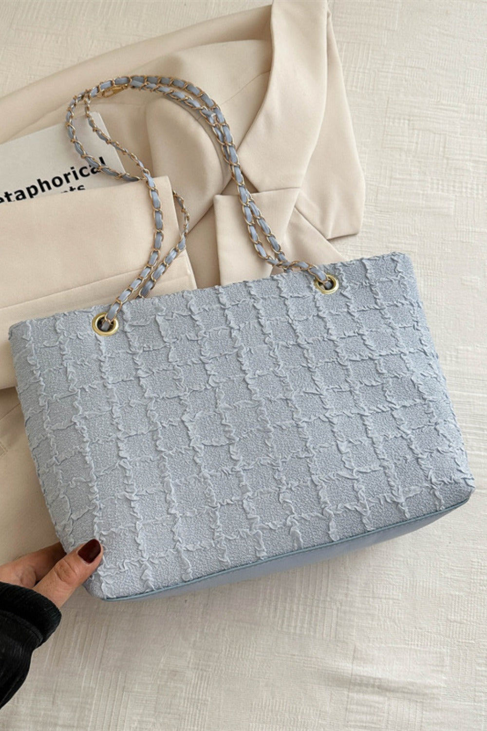 A medium light blue polyester texture tote features a softly structured rectangular silhouette, linked chain handles, and gold-tone hardware as a hand holds its corner.