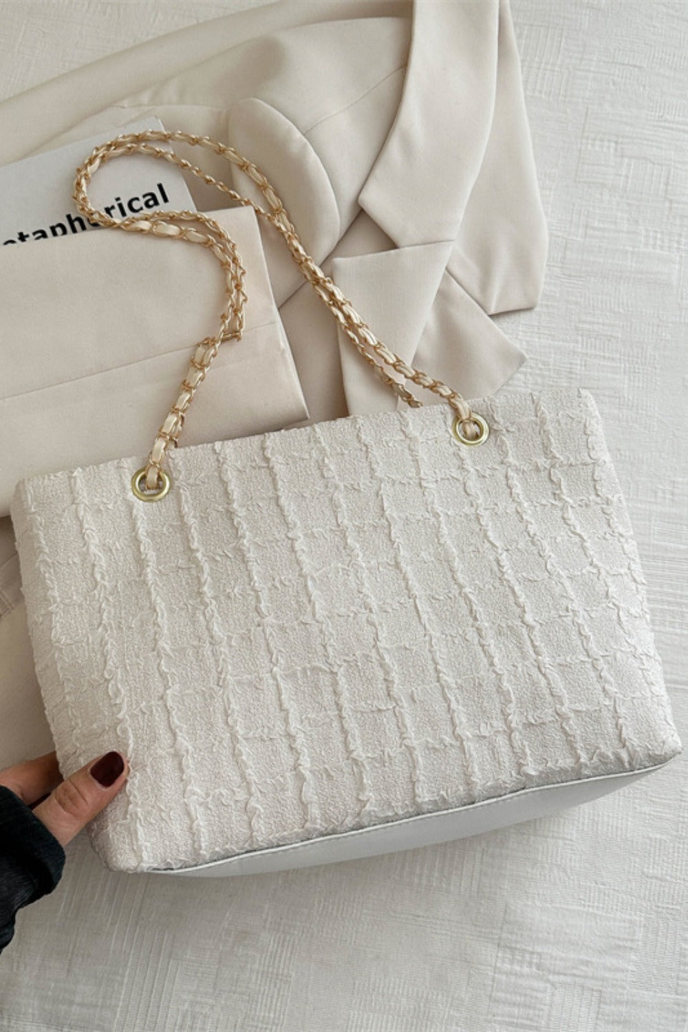 A white medium polyester texture tote features a softly structured silhouette, tactile raised surface, and linked gold-tone chain handles, held at the base by a hand.