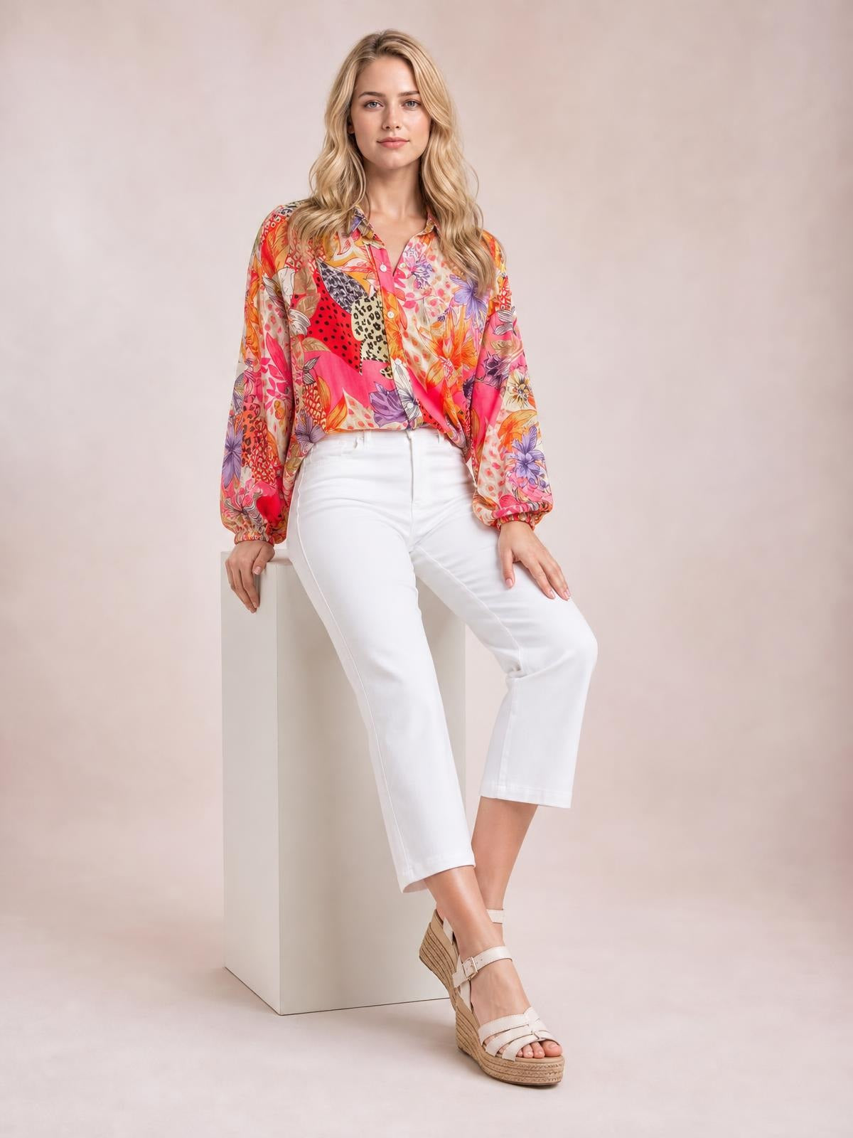 Model seated and facing forward in a colorful printed button-down long-sleeve shirt with a relaxed silhouette, collared neckline, and gathered cuffs.