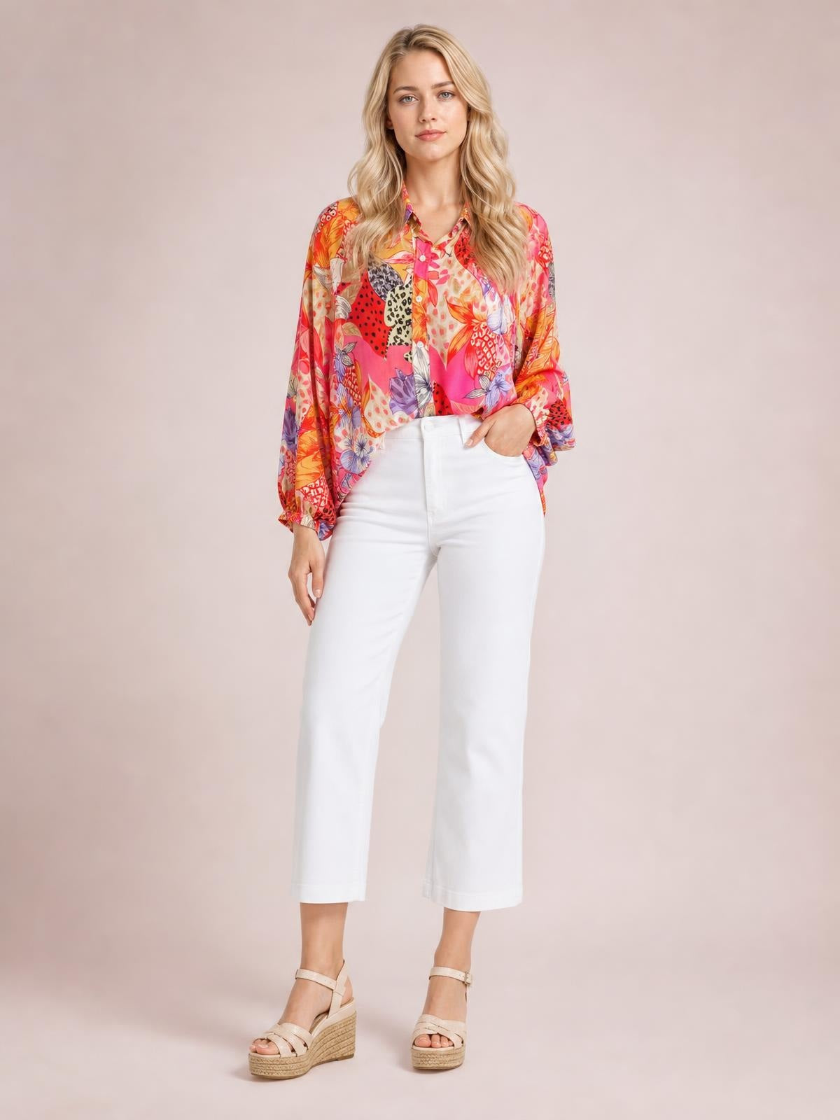 A model stands wearing a vibrant printed button-down shirt with a collared neckline, long sleeves, and a relaxed silhouette in colorful floral and animal motifs.