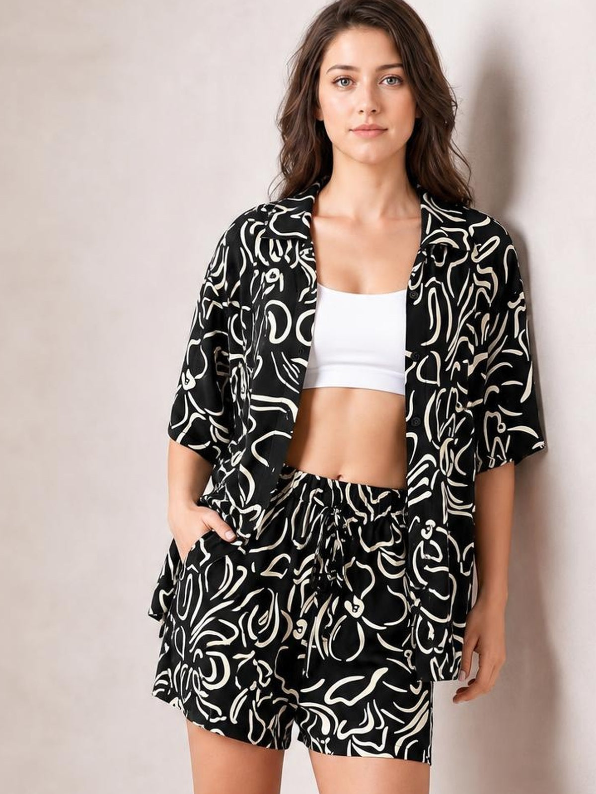 Model poses in a black-and-white printed button-up shirt and shorts set with a relaxed silhouette, short sleeves, pockets, and an elastic waistband.