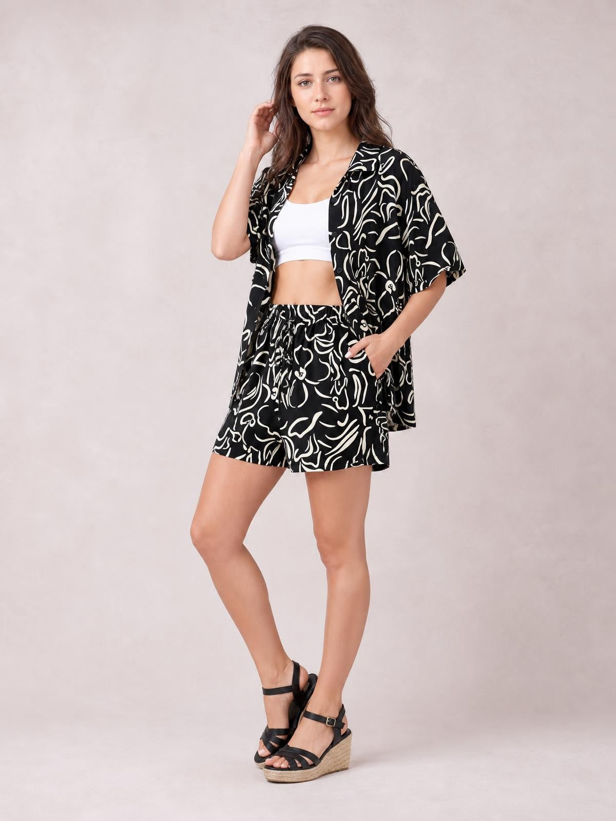 A model poses with one hand in the pocket, wearing a black-and-white printed button-up shirt and matching relaxed shorts with short sleeves and a loose silhouette.