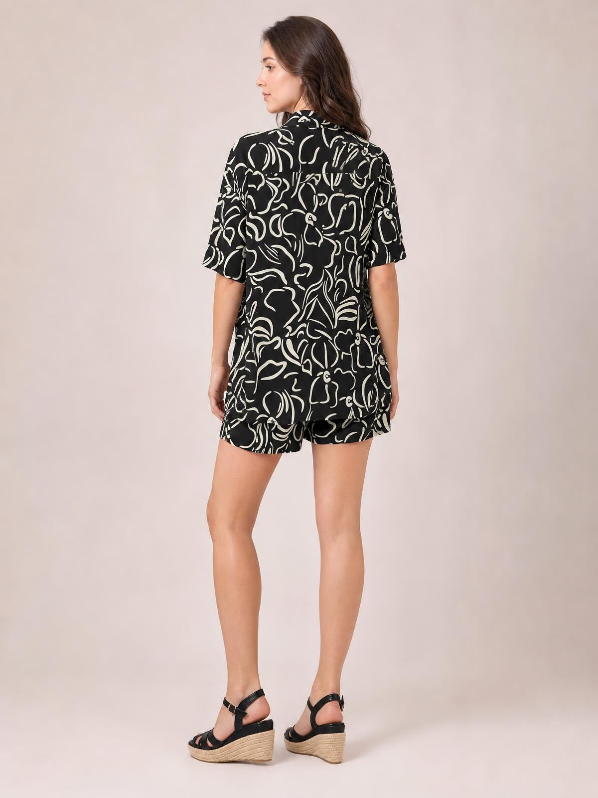 Model turned away in a black printed button-up shirt and shorts set with a relaxed silhouette, short sleeves, collared shirt, and matching shorts.