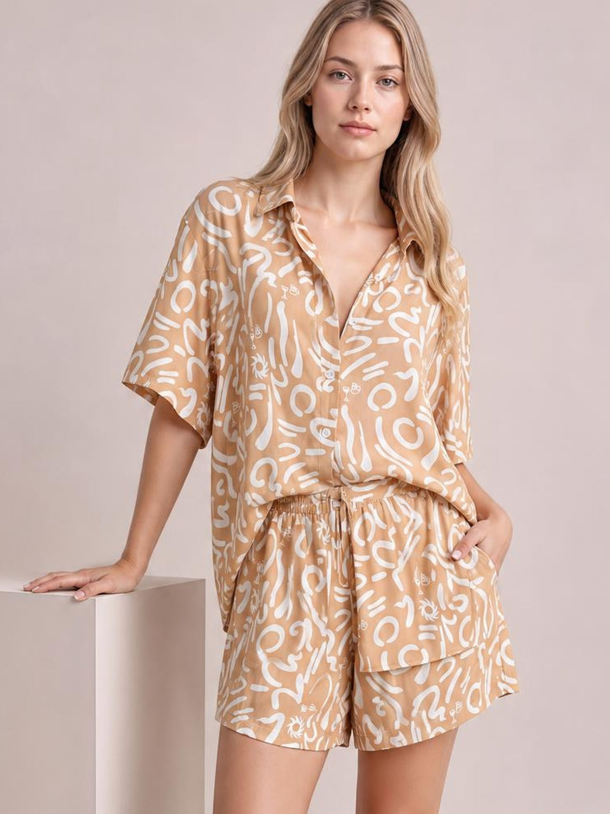 Model poses in a tan printed button-up shirt and matching shorts set with white abstract pattern, short sleeves, relaxed fit, and elastic waist.