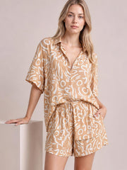 Model poses in a tan printed button-up shirt and matching shorts set with white abstract pattern, short sleeves, relaxed fit, and elastic waist.