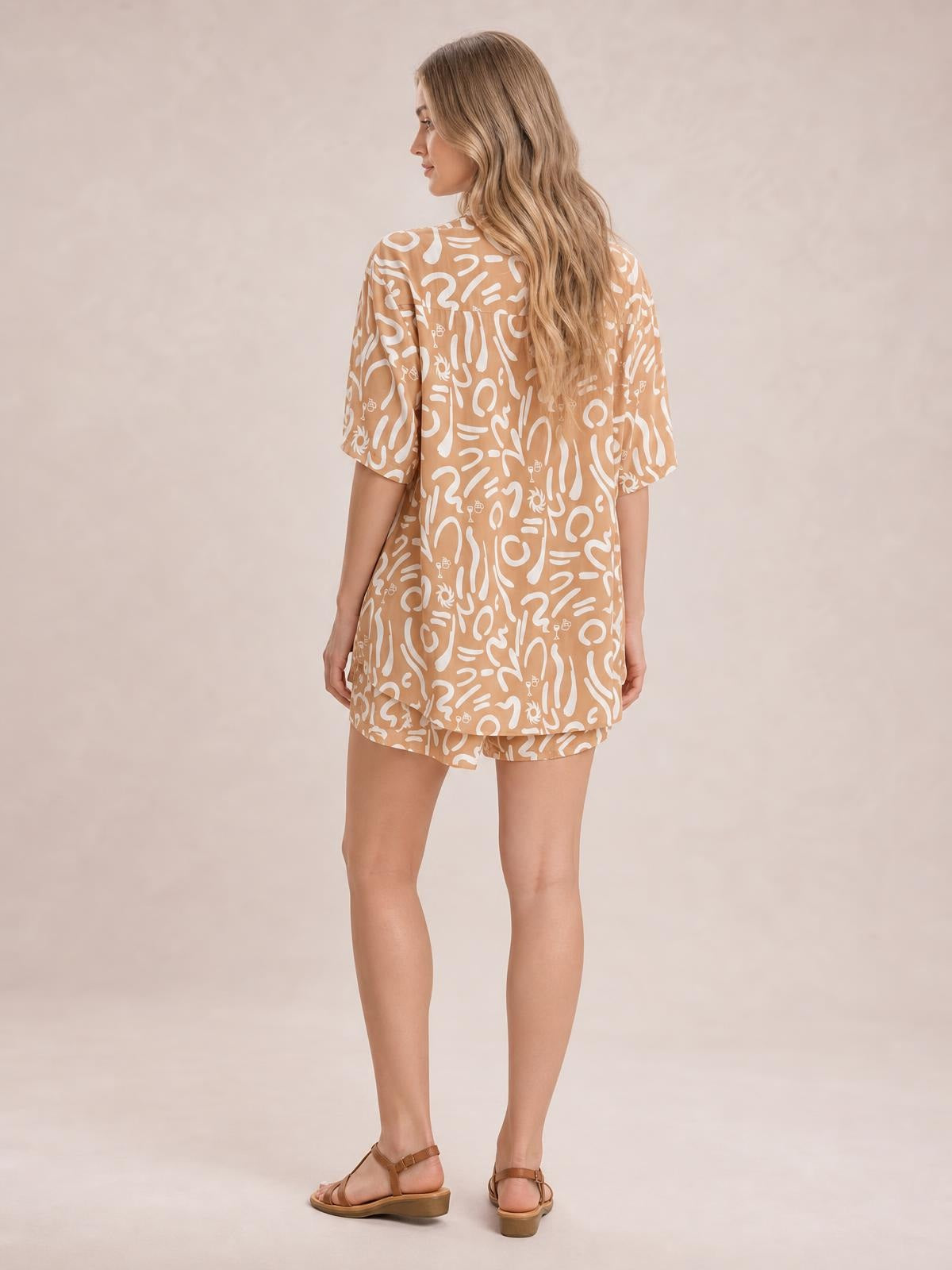 A model poses from the back in a tan printed button-up shirt and matching shorts set, featuring a relaxed short-sleeve silhouette and loose construction.