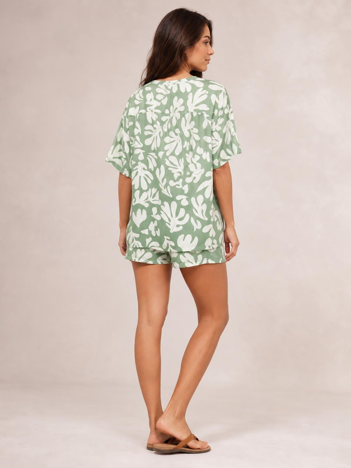 A woman models a sage green and white printed button-up shirt and matching shorts set, featuring a relaxed short-sleeve silhouette as she poses with crossed legs.