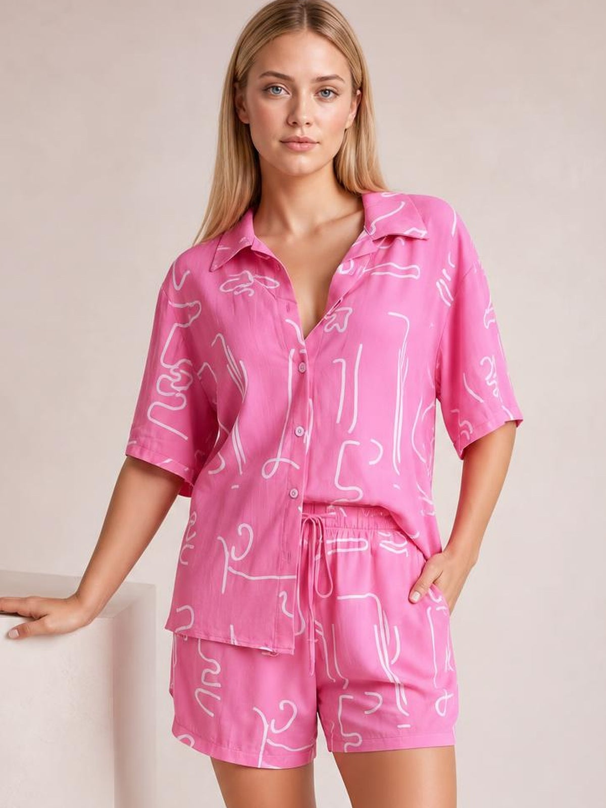 Model poses in a pink printed button-up shirt and shorts set with white abstract lines, short sleeves, collared shirt, and drawstring waist.