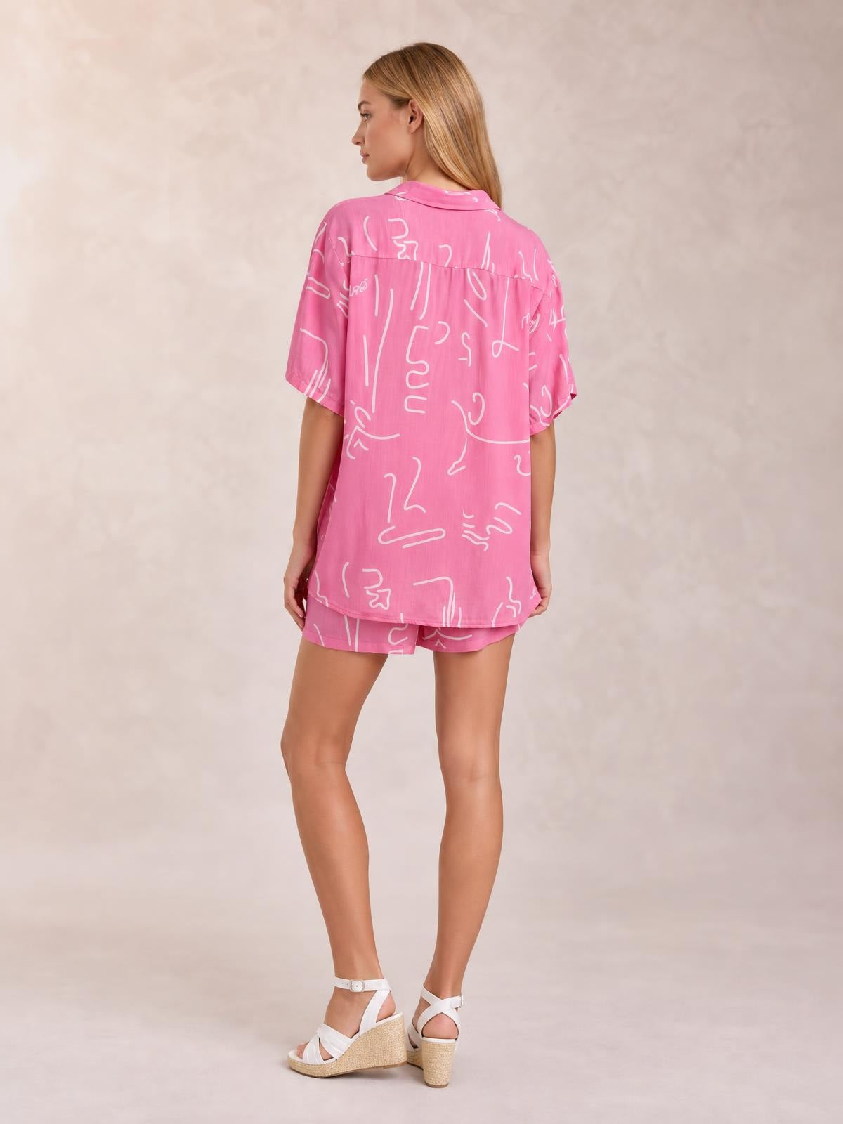 Model poses in a pink printed button-up shirt and shorts set, shown from behind with a relaxed loose silhouette, short sleeves, and collared construction.