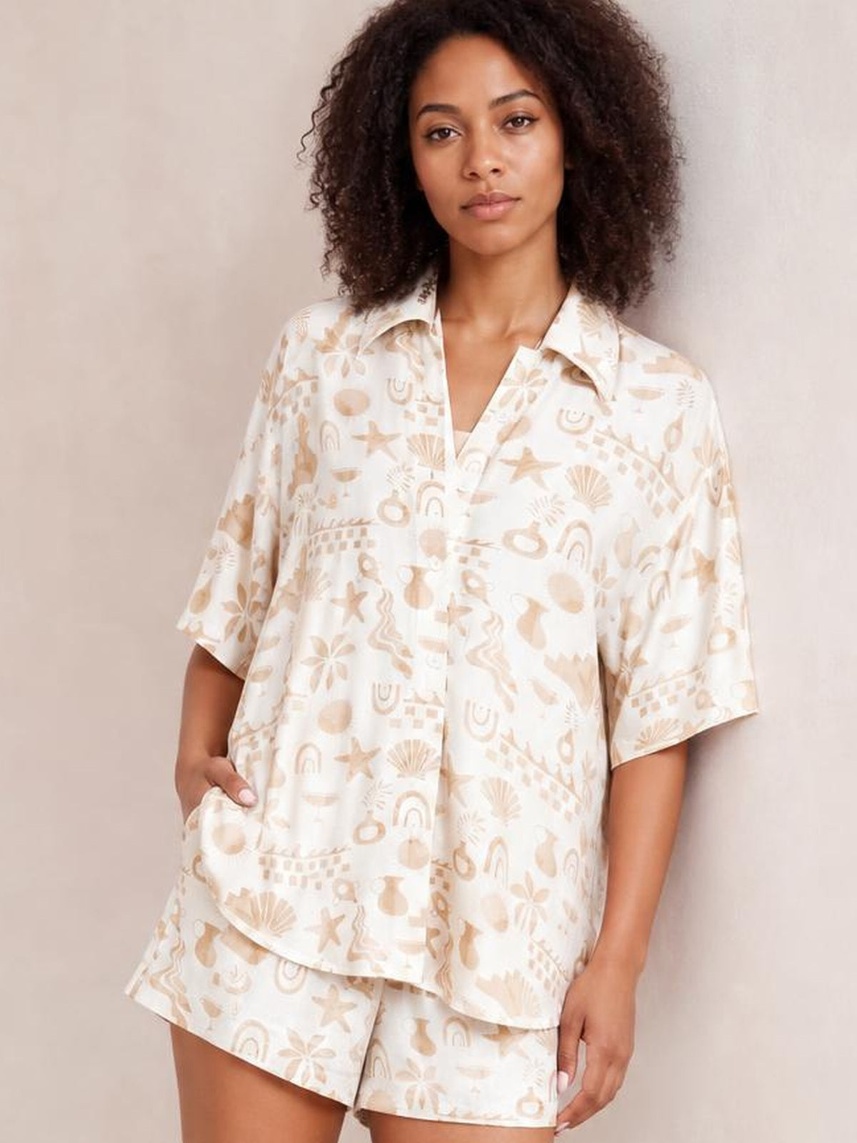 Model poses in a cream printed button-up shirt and matching shorts set, featuring beige motifs, short sleeves, a relaxed silhouette, and a hand in the pocket.
