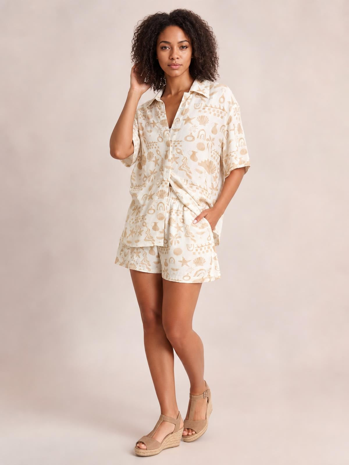 A model poses in a cream printed button-up shirt and matching shorts set with a relaxed silhouette, short sleeves, button front, and pockets.