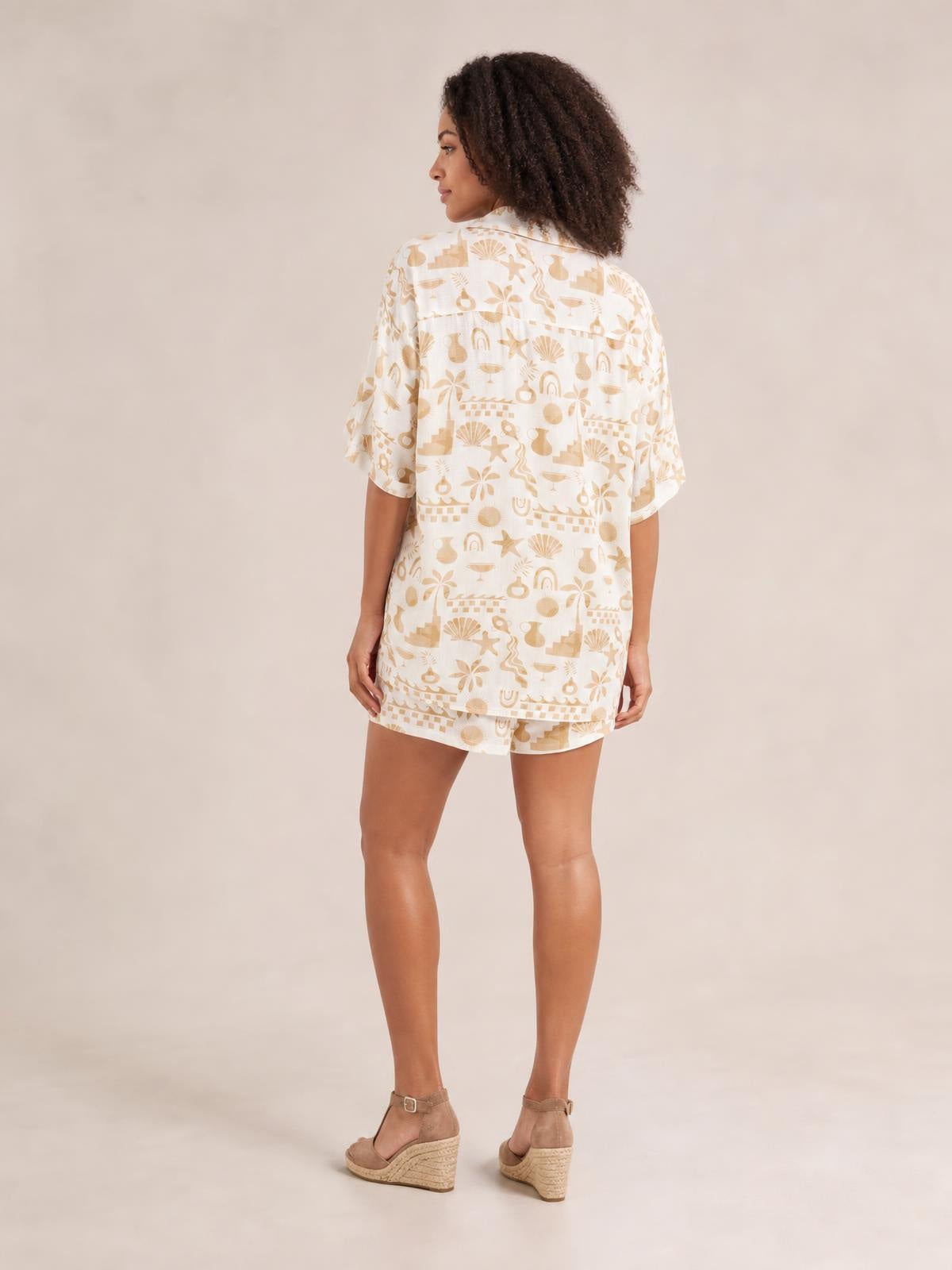 Model stands in a three-quarter back pose wearing a cream printed button-up shirt and matching shorts set with a relaxed silhouette and short sleeves.