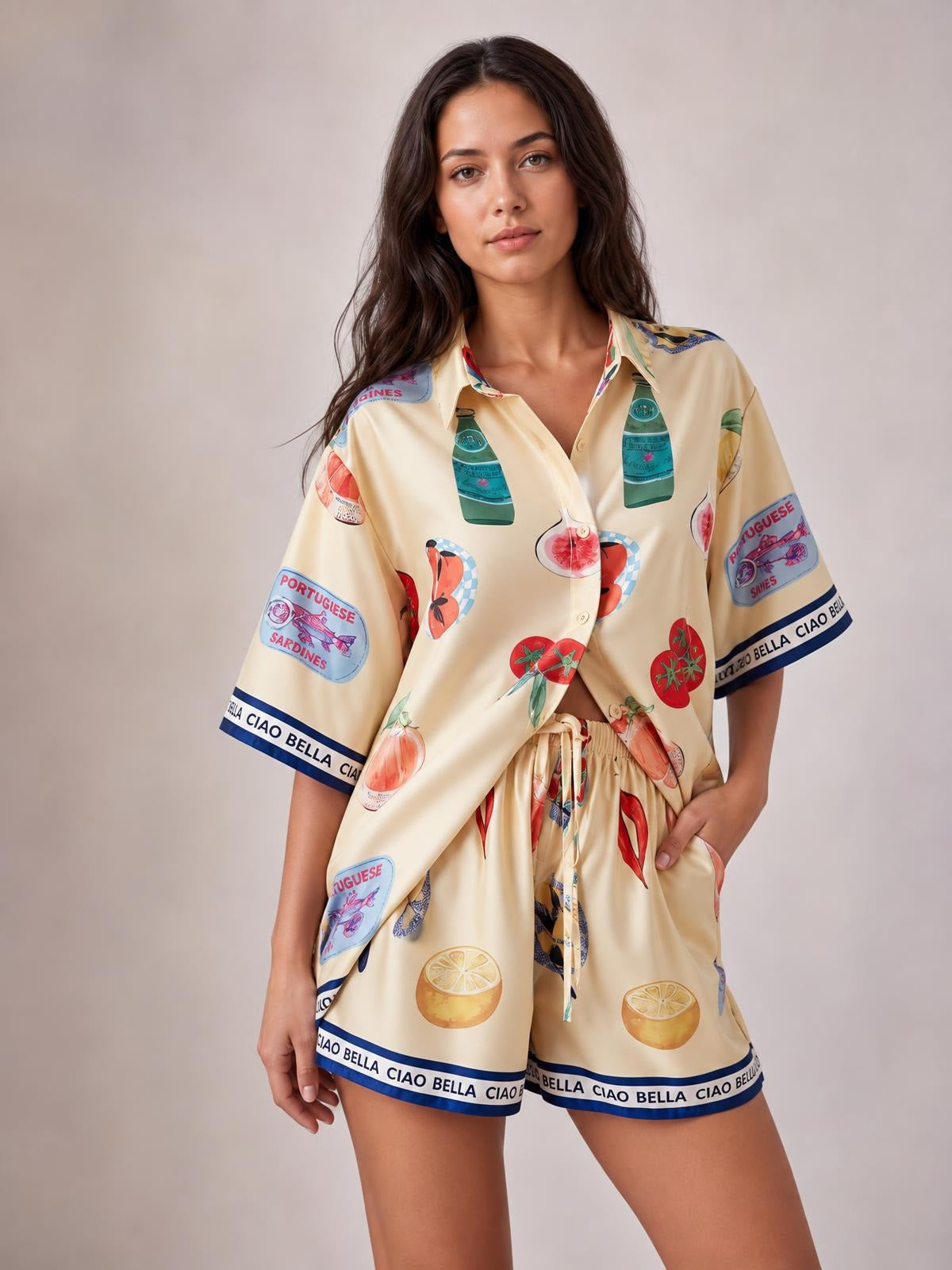 A model wears a relaxed cream printed button-up top with short sleeves and matching drawstring shorts, featuring colorful fruit and bottle motifs with navy borders.