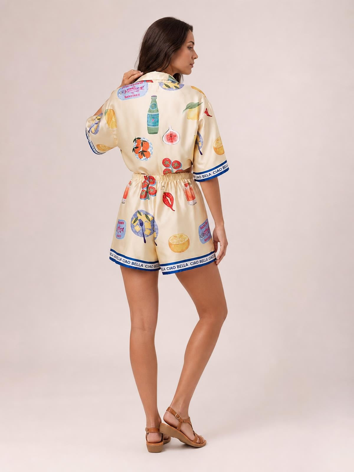 Model turned away in a relaxed cream printed button-up top and matching drawstring shorts, featuring short sleeves, colorful food motifs, and contrast trim.