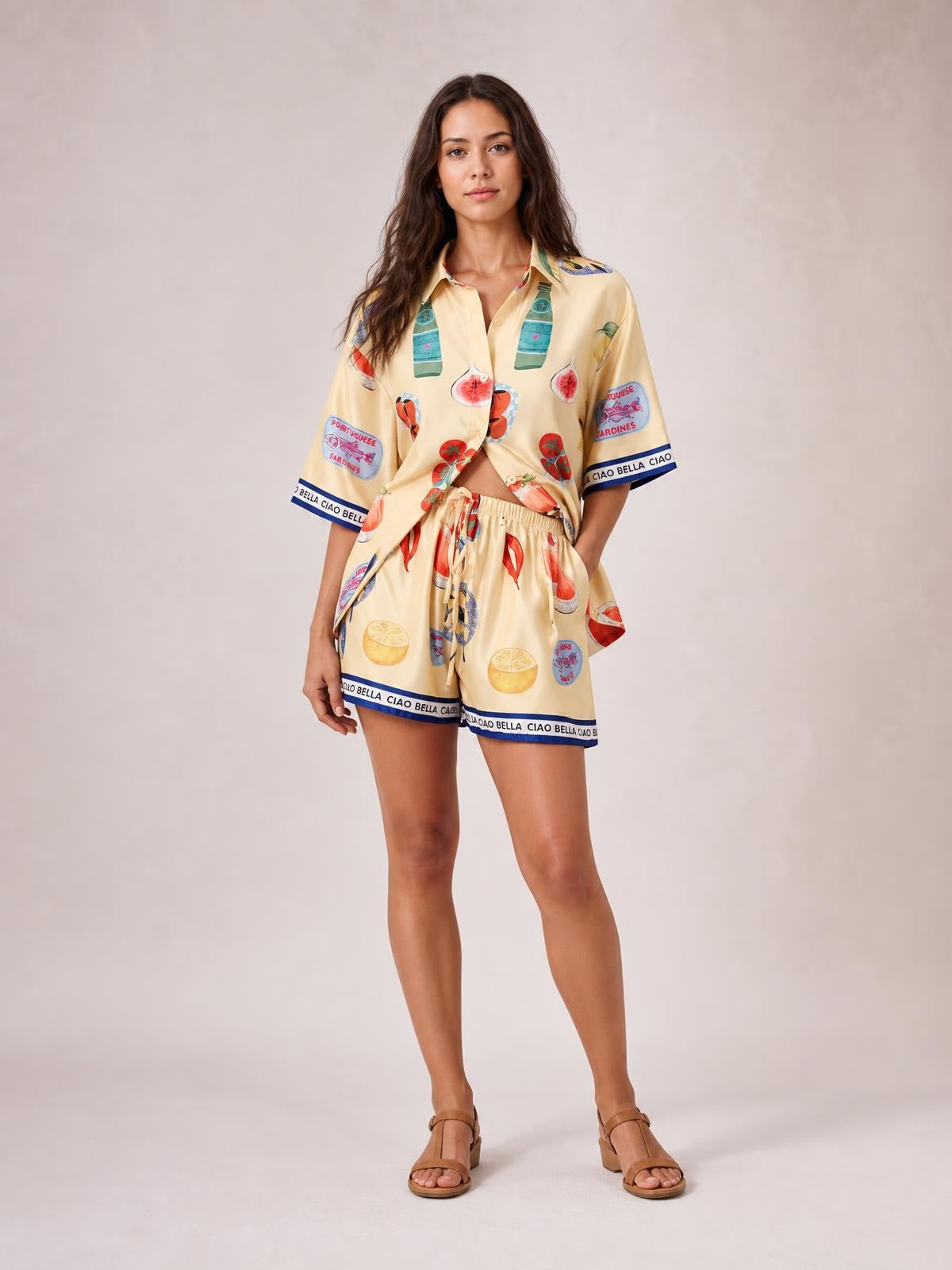 Model wears a relaxed printed button-up top and matching drawstring shorts set in pale yellow, with short sleeves, blue trim, and a straight silhouette.