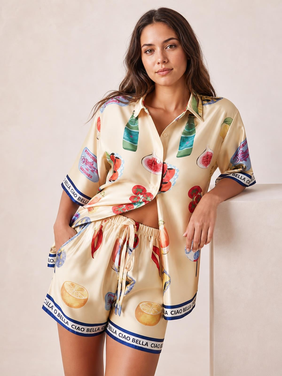 Model wears a relaxed cream printed button-up top with short sleeves and matching drawstring shorts, featuring colorful fruit and border graphics in a standing pose.